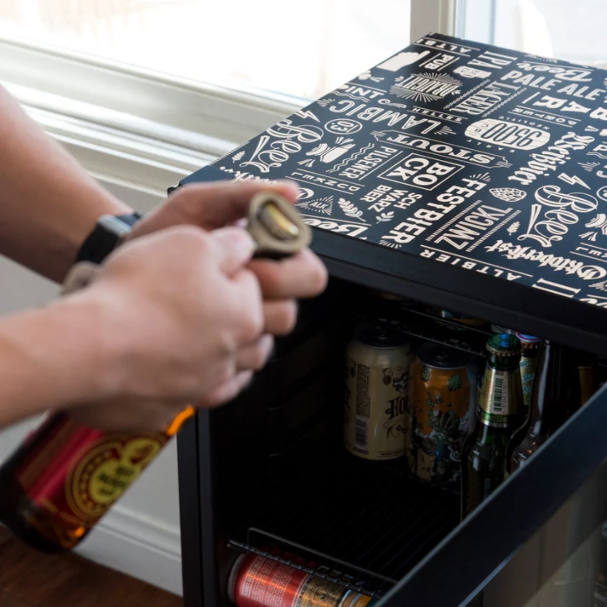 Newair 126-Can Freestanding Beers of the World Fridge with SplitShelf - Man opening a bottle beside a beer-themed fridge