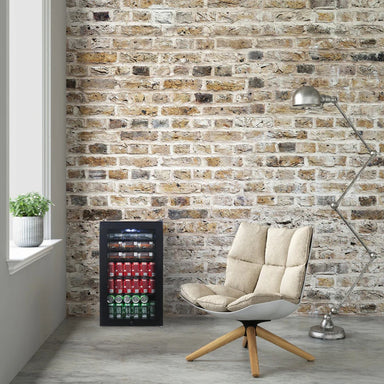 Newair 126-Can Freestanding Beverage Fridge – 19″ Black - fridge against a brick wall beside a modern cushioned chair and floor lamp