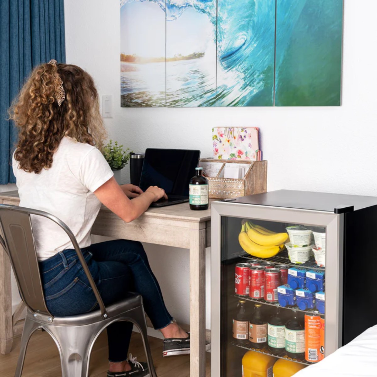 Newair 126 Can Freestanding Beverage Fridge – 18.9" Stainless Steel - Woman working at a desk beside the fridge stocked with drinks and snacks