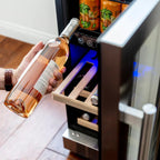Newair 15″ SplitShelf 48-Can / 9-Bottle Dual Zone Beverage Fridge - Person holding a wine bottle while pulling out a wooden shelf from a stainless steel beverage fridge